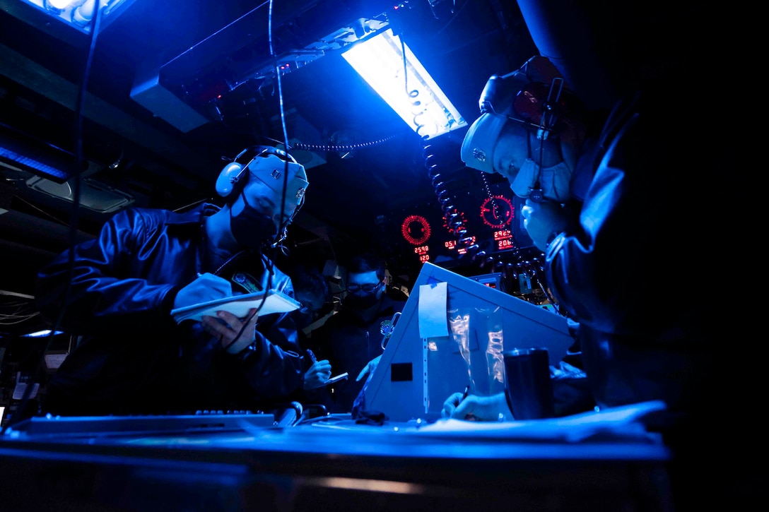 Two sailors illuminated by blue light wear headphones while one writes on a notepad.