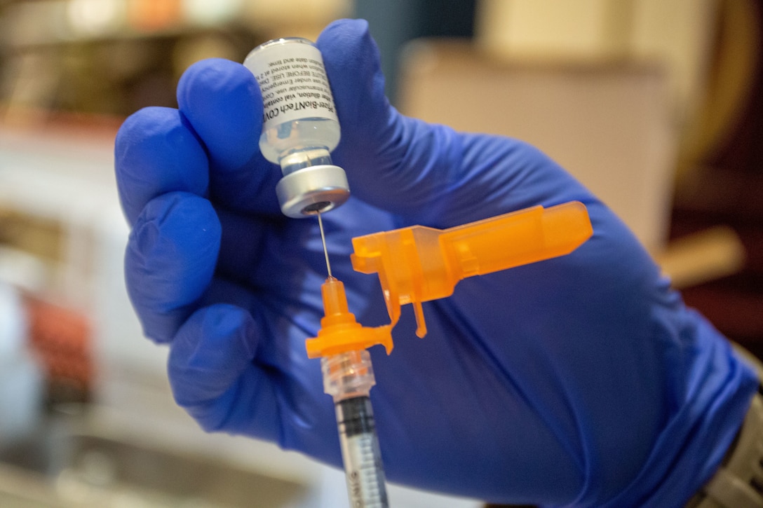 A gloved hand holds a needle that draws a vaccine from a vial.