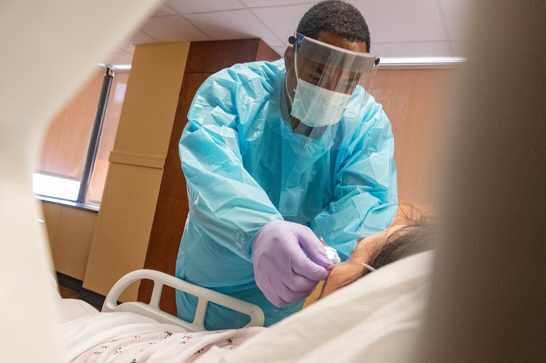 An airman in medical and protective gear stands above a patient in a hospital bed.