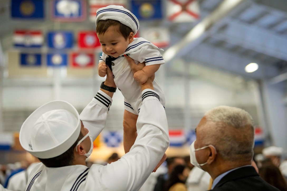 A sailor lifts his son in the air while standing next to his father at a graduation ceremony.