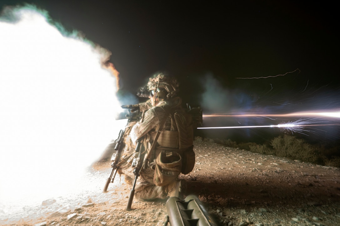 Marines kneel while firing a weapon in the dark illuminated by bright striking lights.