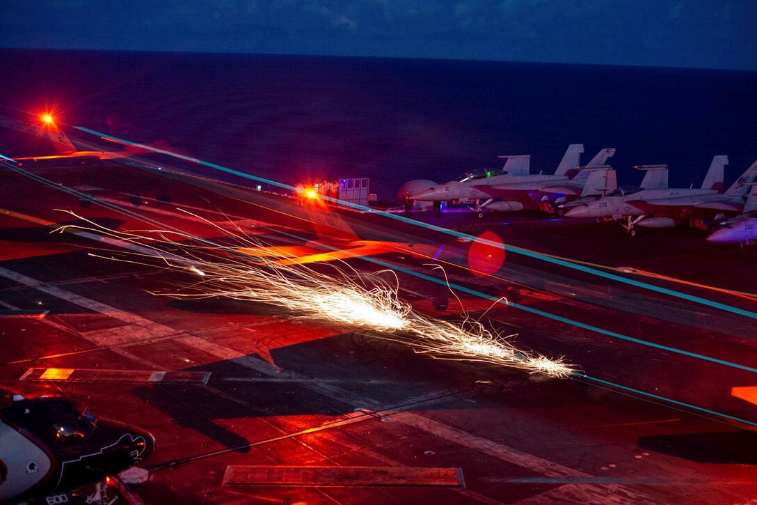 An aircraft lands on a ship illuminated by colorful lights.