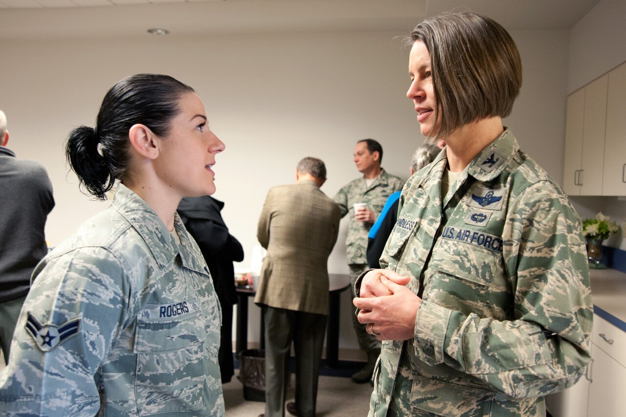 Two female airmen talk to each other in a room with others in the background.