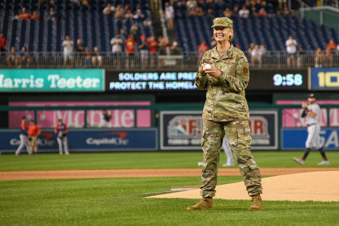 A woman in uniform and holding a baseball stands near a pitchers mound in a stadium.