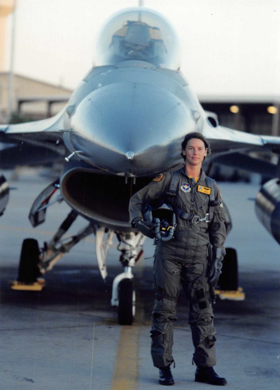 A woman in a pilot's uniform and gear holds a helmet under her arm in front of a fighter jet.
