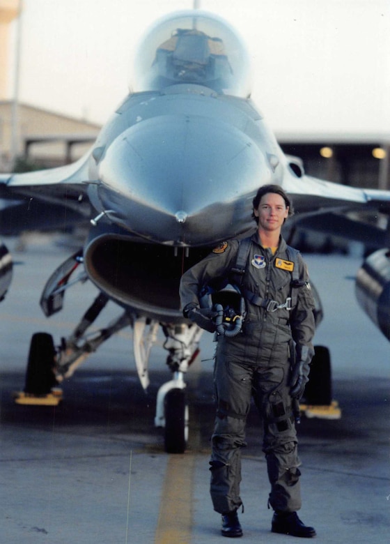 A woman in a pilot's uniform and gear holds a helmet under her arm in front of a fighter jet.