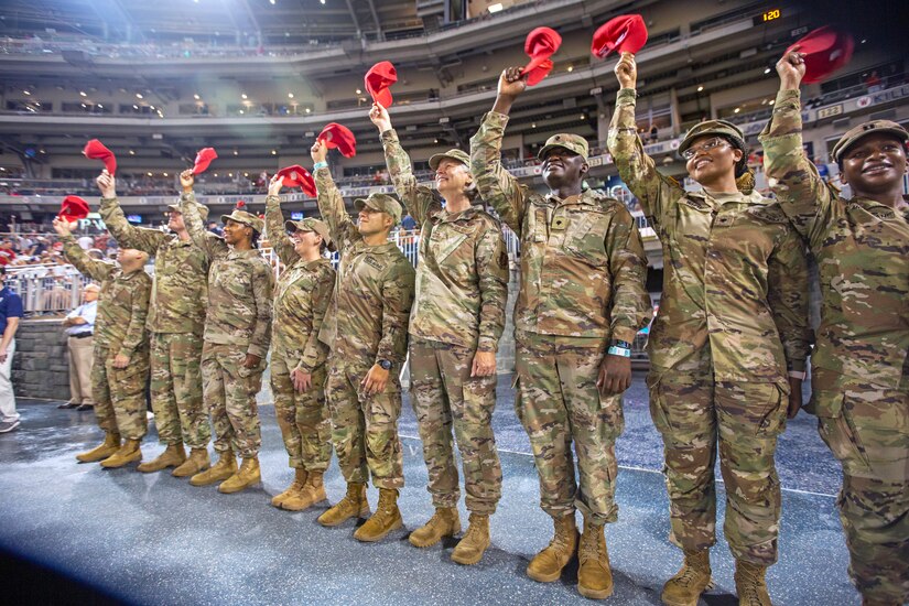 Nine service members in uniform hold red hats high above their heads to salute people in a stadium.