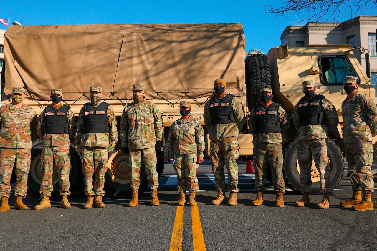 Nine service members in uniform, some of whom wear vests, pose in front of a large military truck.
