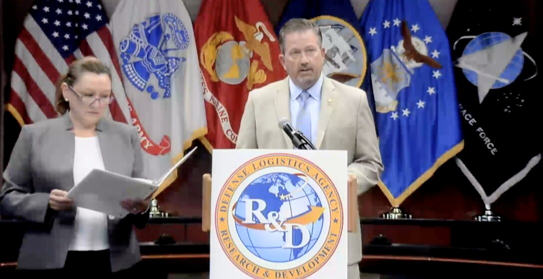A man stands at a podium with the US, DLA and service flags behind him and a woman slightly behind him with a folder in her hands.