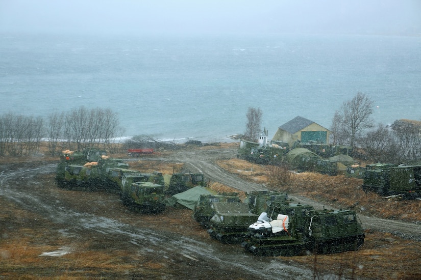 Military vehicles are parked in a muddy area along a coastline.