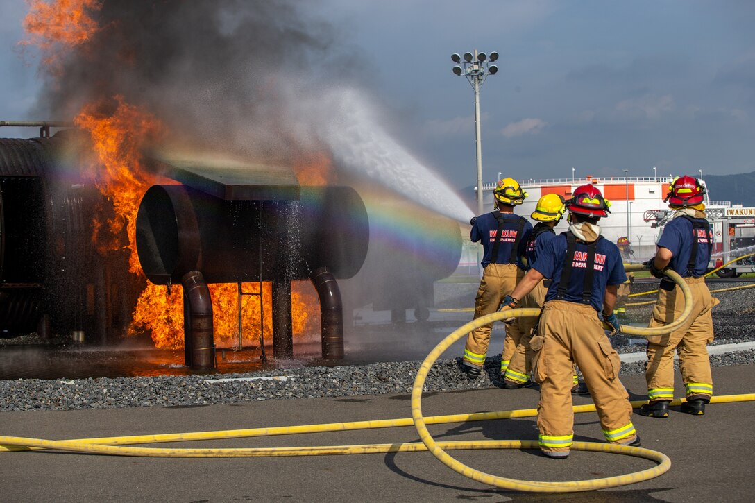Marines in firefighting gear spray water on a fire.
