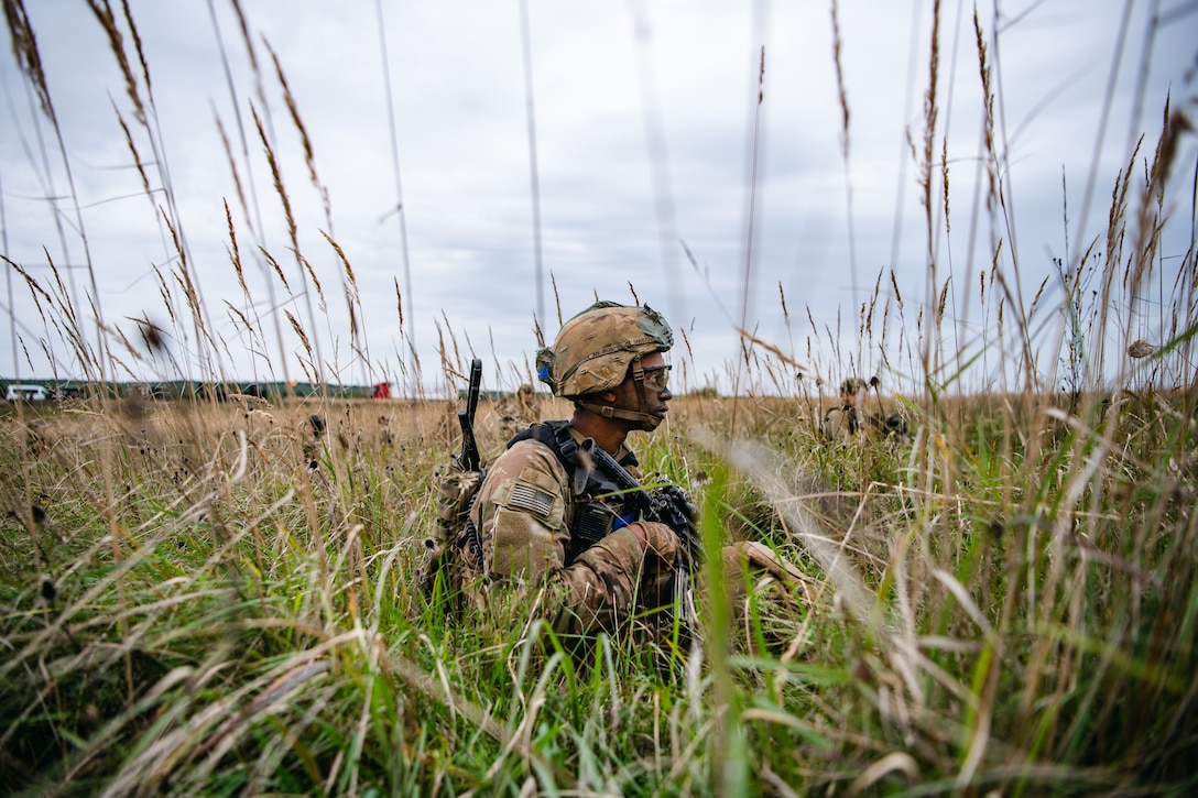 A paratrooper holding a weapon walks through a field of tall grass.