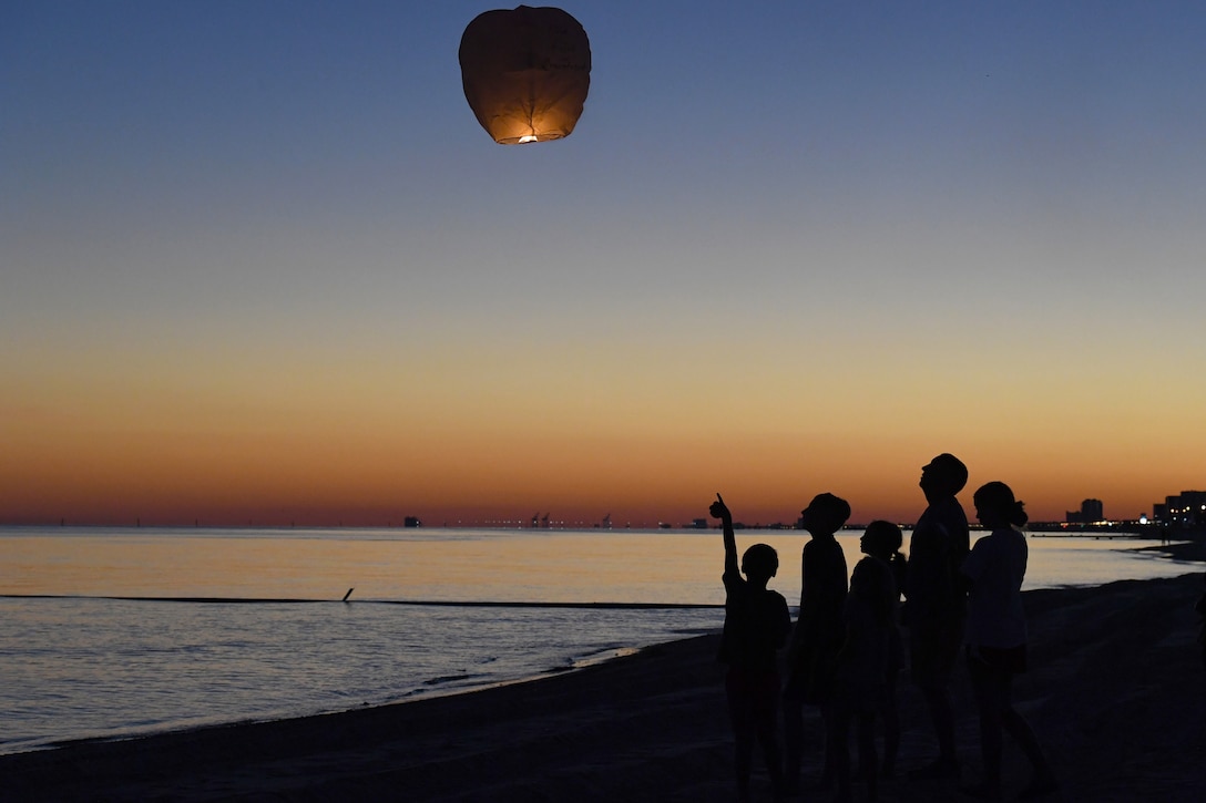 A group of people stand on a beach at twilight as a lantern balloon floats in the sky.