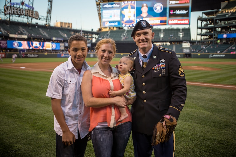 A soldier in dress uniform poses with a woman, a boy and a toddler on a baseball field.