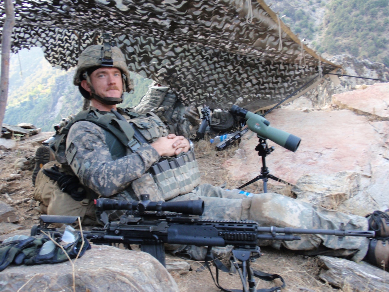 A soldier in combat gear leans against a cargo pack beside a scope and a rifle.