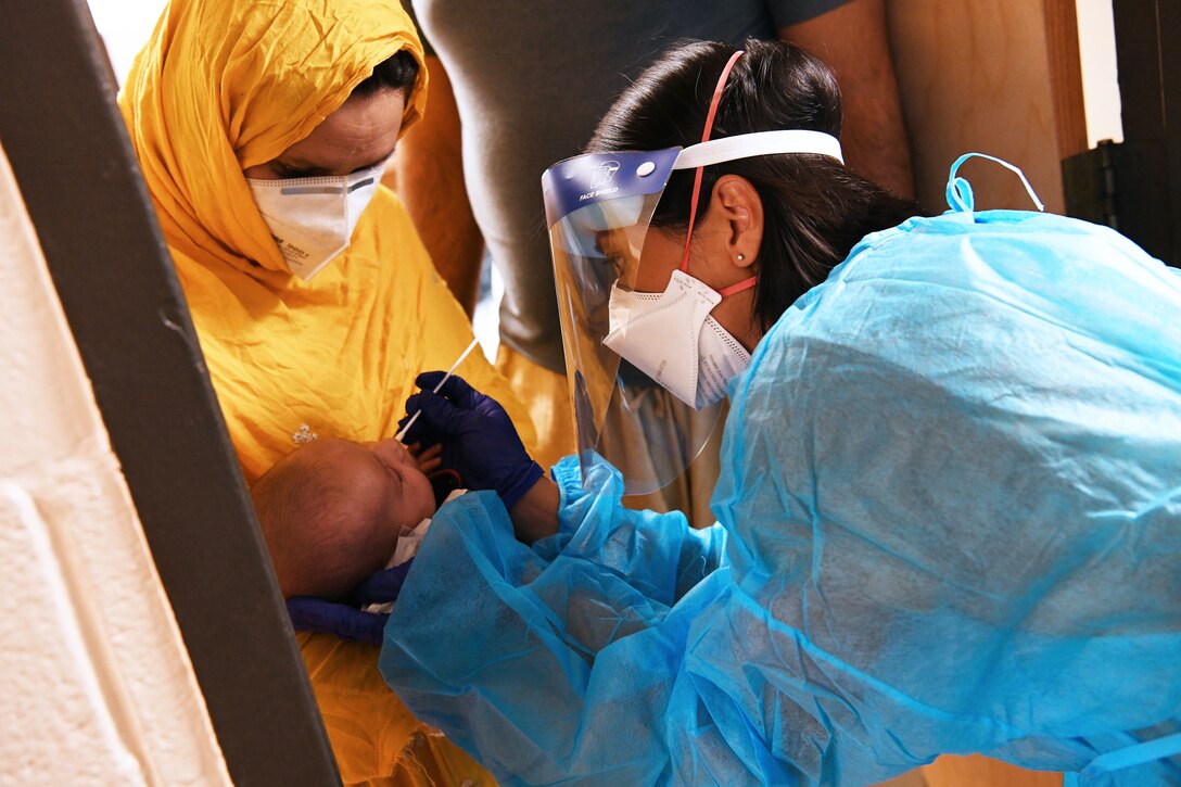 A female soldier wearing personal protective equipment holds a nasal swab to the nose of an infant; the child is being held by a woman who's also wearing a face mask.