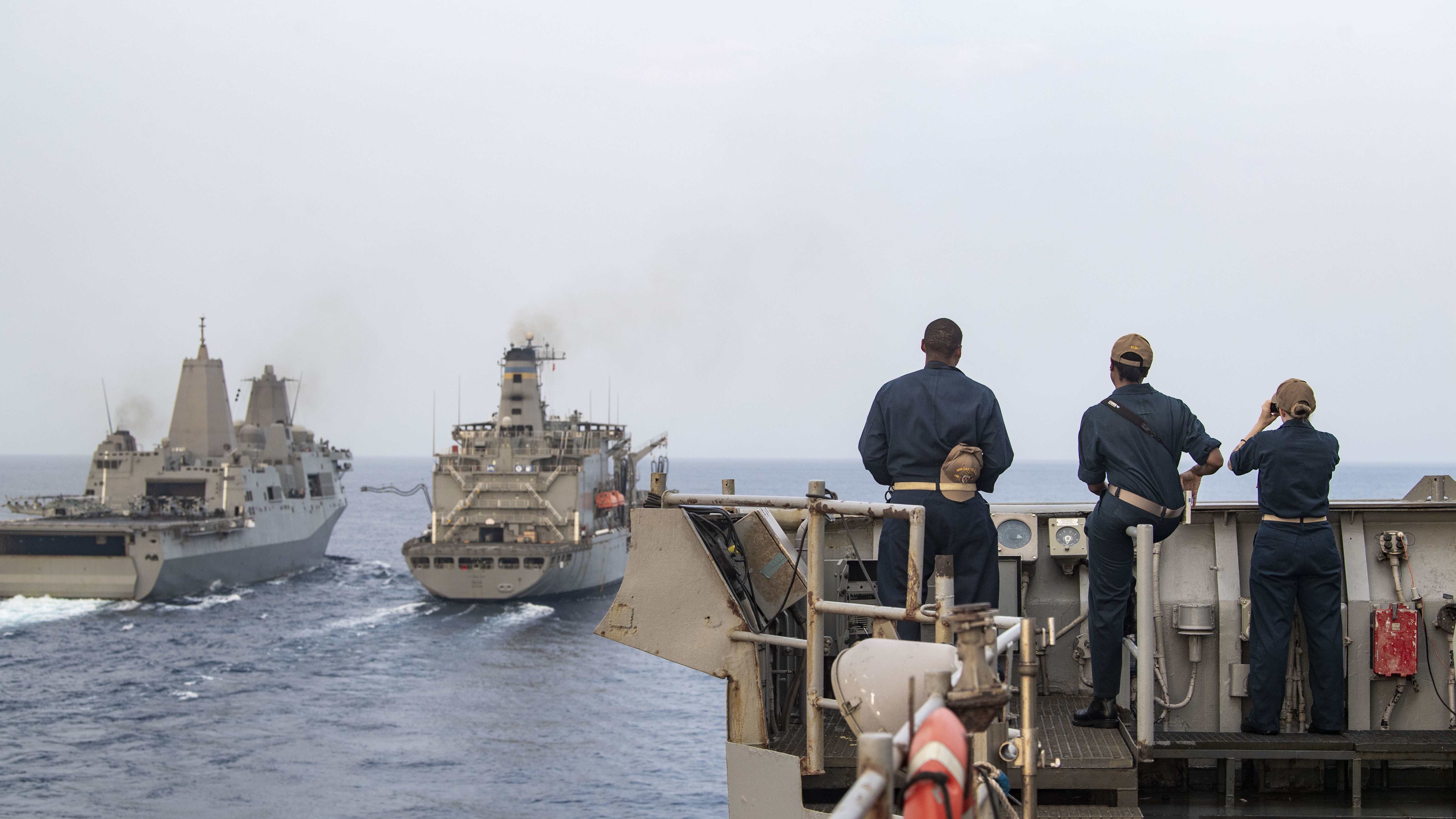 Sailors look on during a replenishment-at-sea with the Henry J. Kaiser-class underway replenishment oiler USNS Pecos (T-AO 197), right, and the amphibious transport dock ship USS San Antonio (LPD 17).
