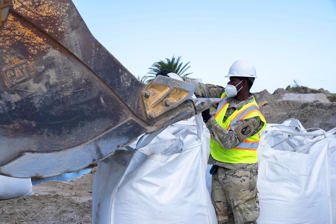 A soldier hooks straps of a 3-by-3 foot sack to an excavator bucket.