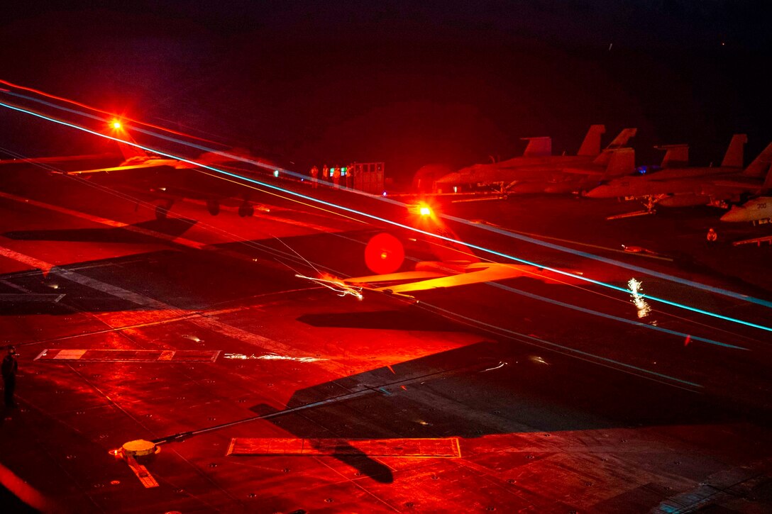 Aircraft illuminated by red lights recovers on the deck of a ship.