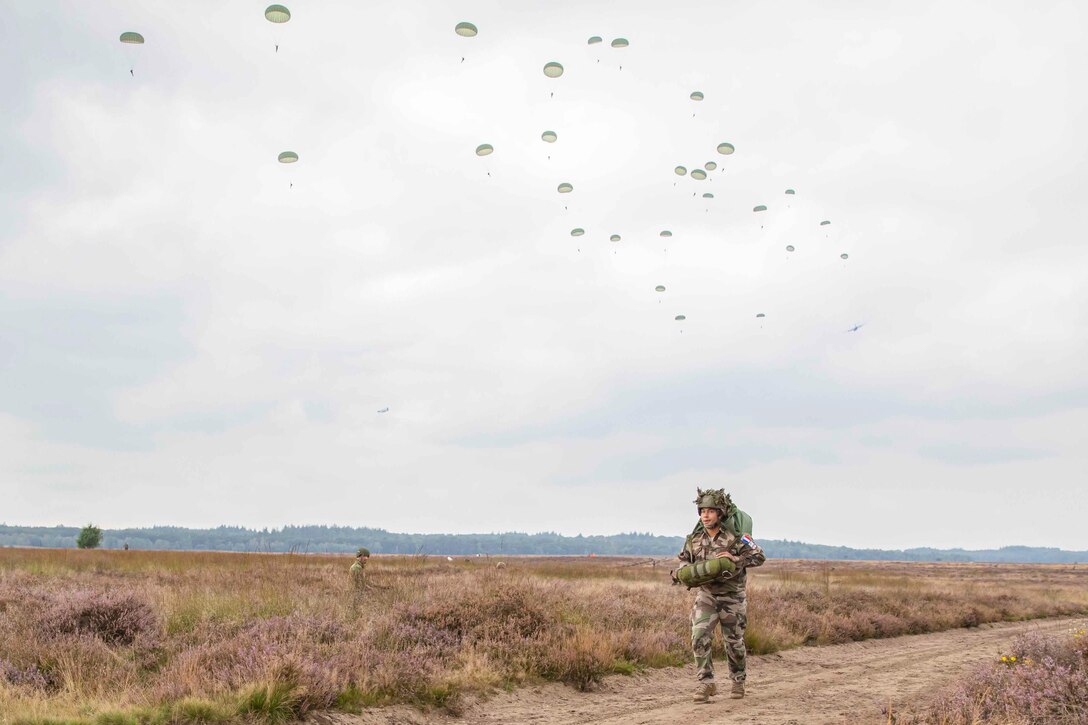 A soldier walks alongside a dirt road as others descend in the sky wearing parachutes.