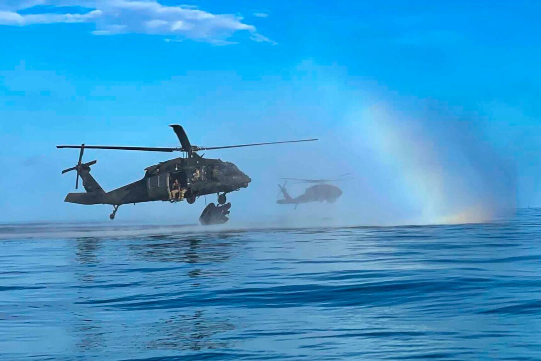 Two helicopters fly over water as a rainbow shines in the background.