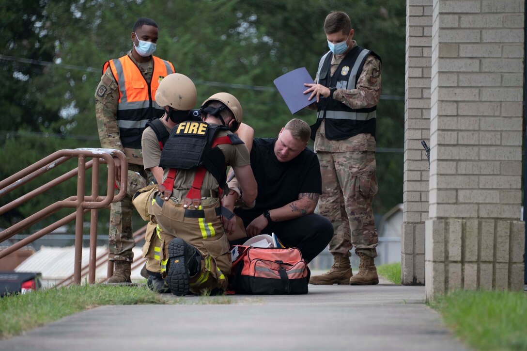 Two emergency responders assist an exercise volunteer victim with moulaged wounds. Two Airmen stand observing the scene.