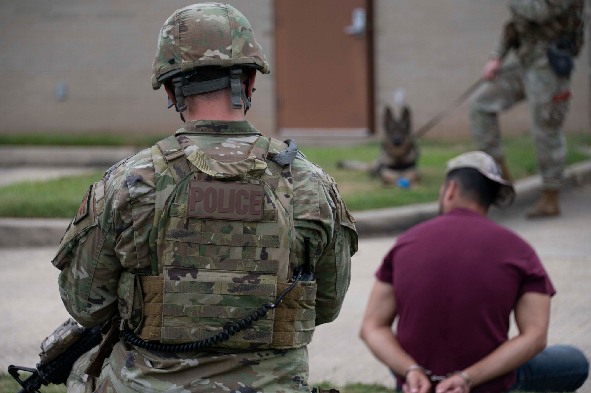 Military police officer and K-9 officer stare at a man sitting in handcuffs.
