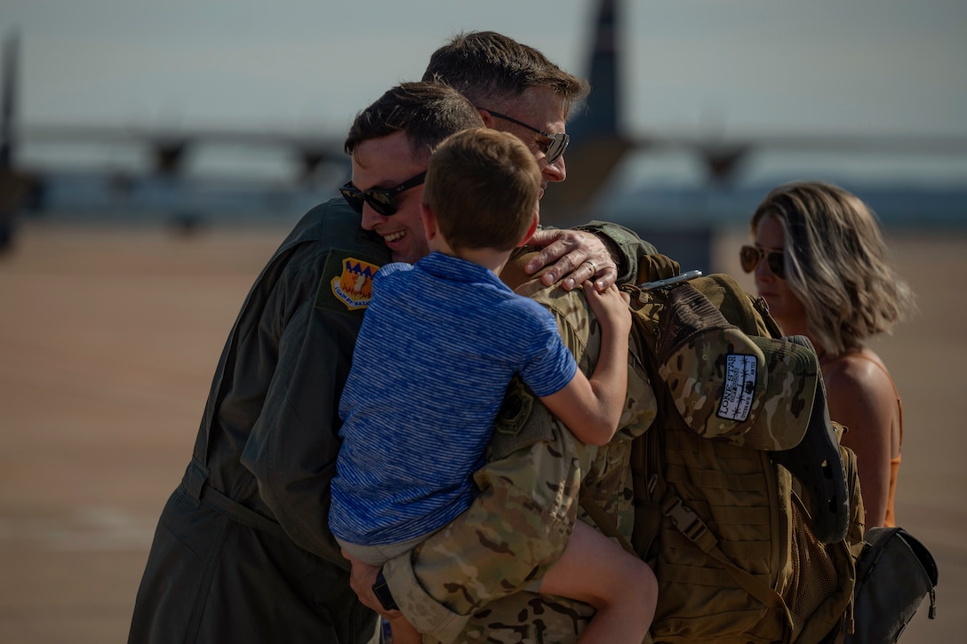 An airman holding a child hugs a fellow airman on a tarmac near aircraft.