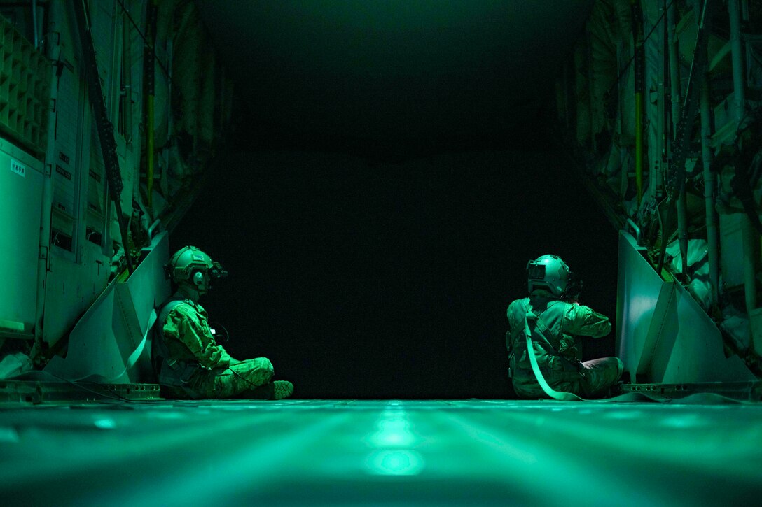 Two airmen illuminated by green light sit at the back of an aircraft at night.