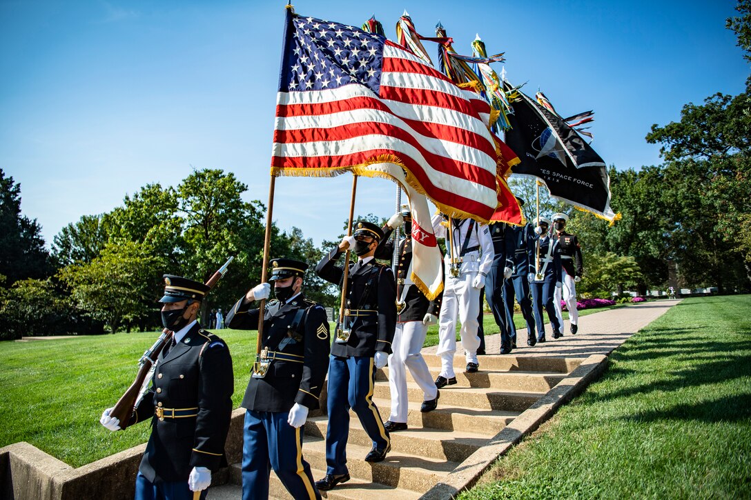 A line of service members in dress uniforms carry flags down some steps.