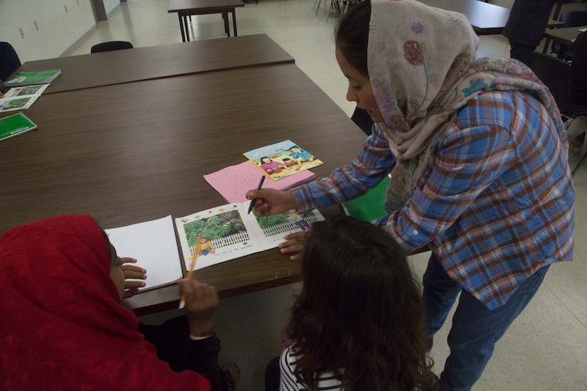 A woman in a headscarf leans over a picture book on a table. Young children watch.