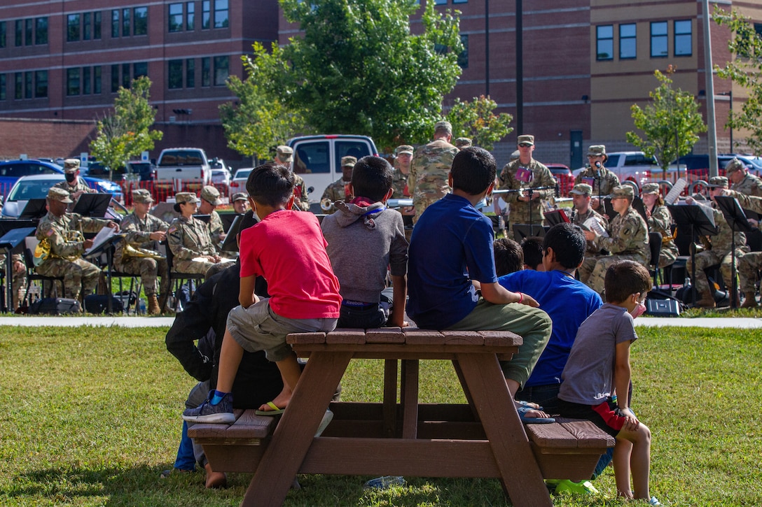 Children sit on a  picnic table. In the background, soldiers play instruments.