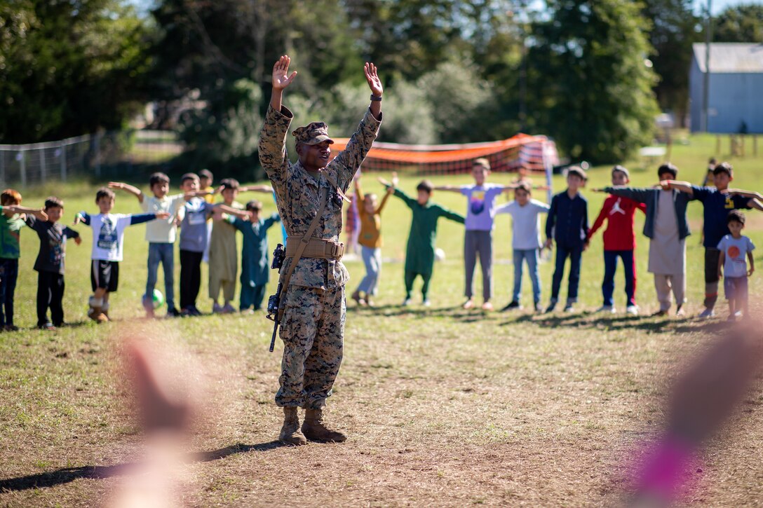 A uniformed service member stands with his arms in the air. Around him stand children mimicking his actions.