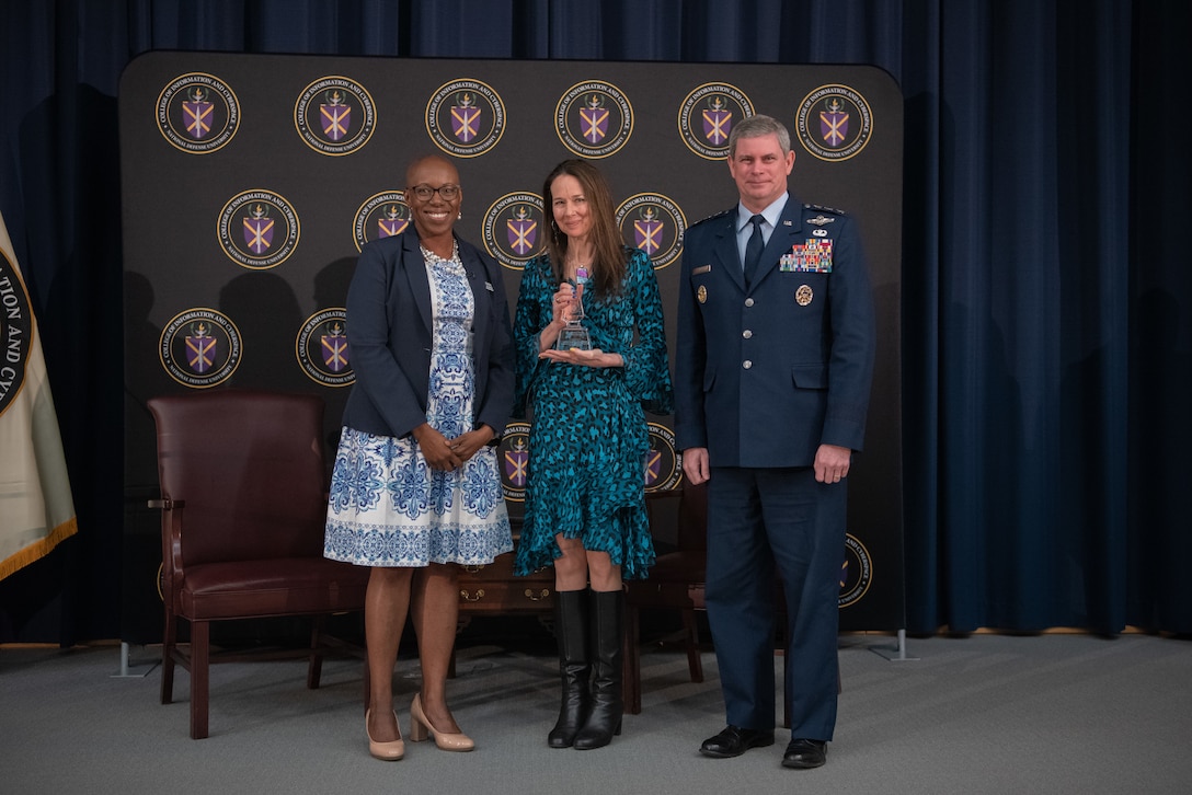 Two women and a man wearing a military uniform stand next to one another. The woman in the center holds an award.