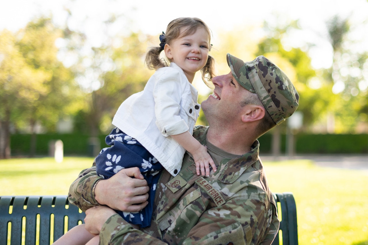 An airman smiles at his daughter while holding her.