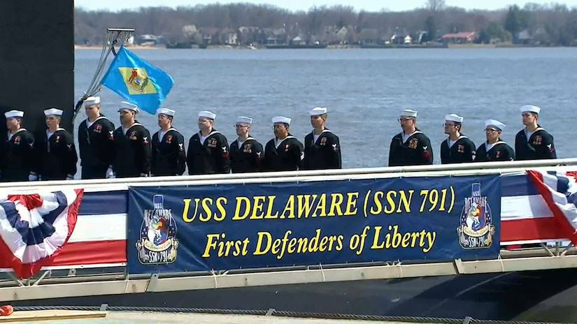 Sailors line up behind a banner bearing the words "USS Delaware".