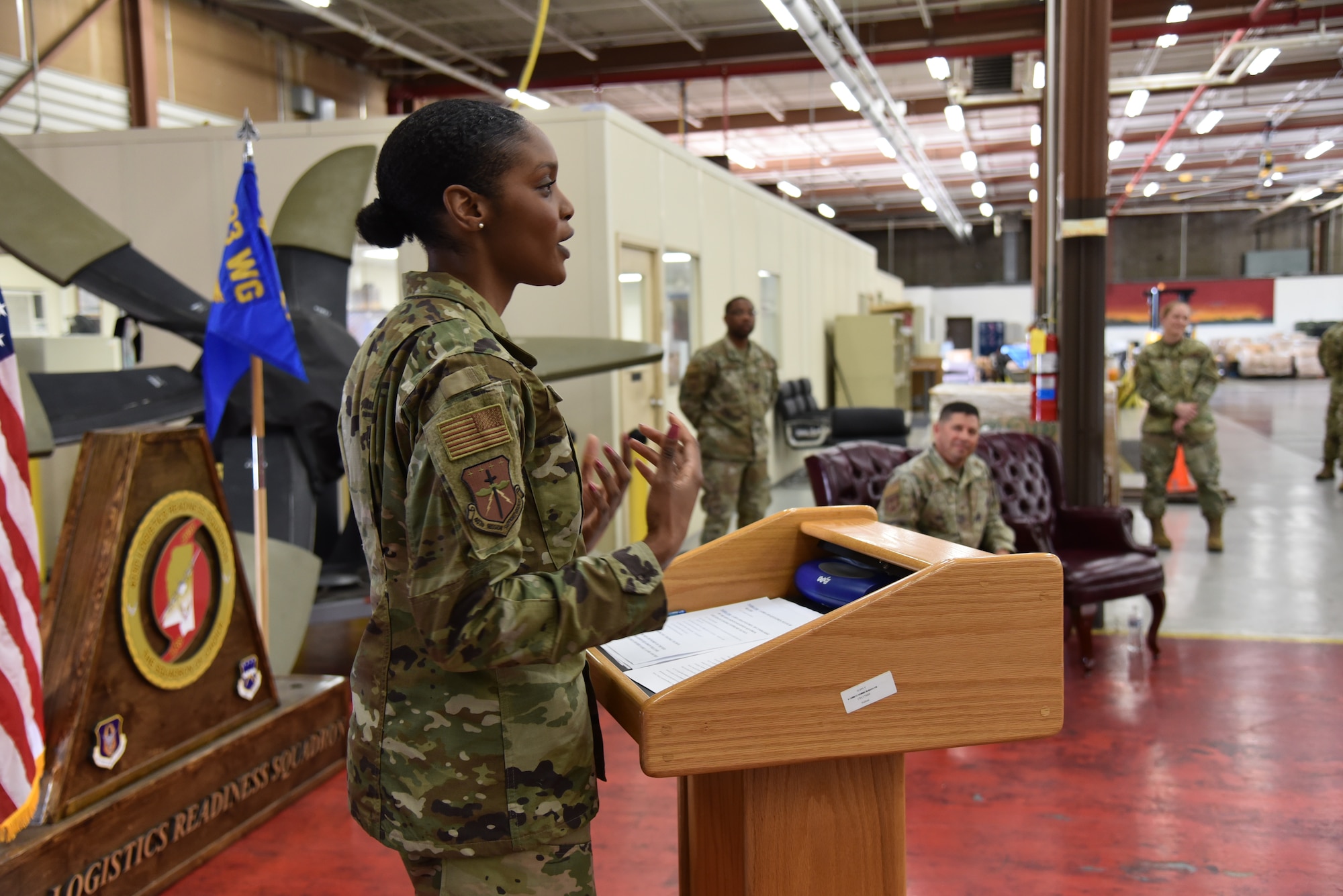 Lt. Col. Tillman stands behind a podium as people in the background listen to her speak