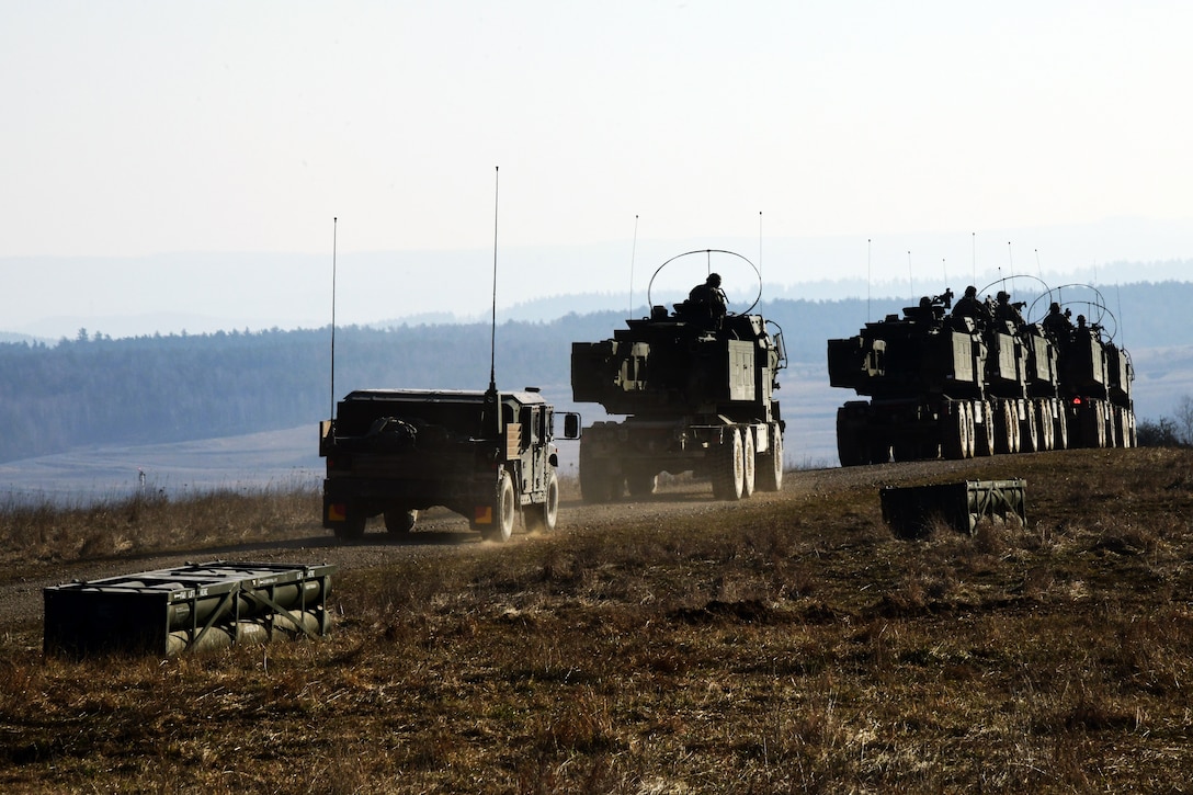 Several trucks of soldiers drive up a road during a live fire exercise.