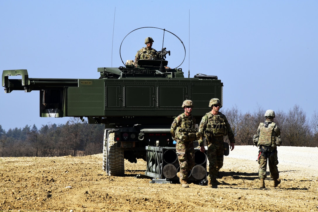 Several soldiers on the ground and one on the truck participate in a live fire exercise.