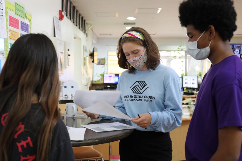 Three teens stand and talk and look at papers.
