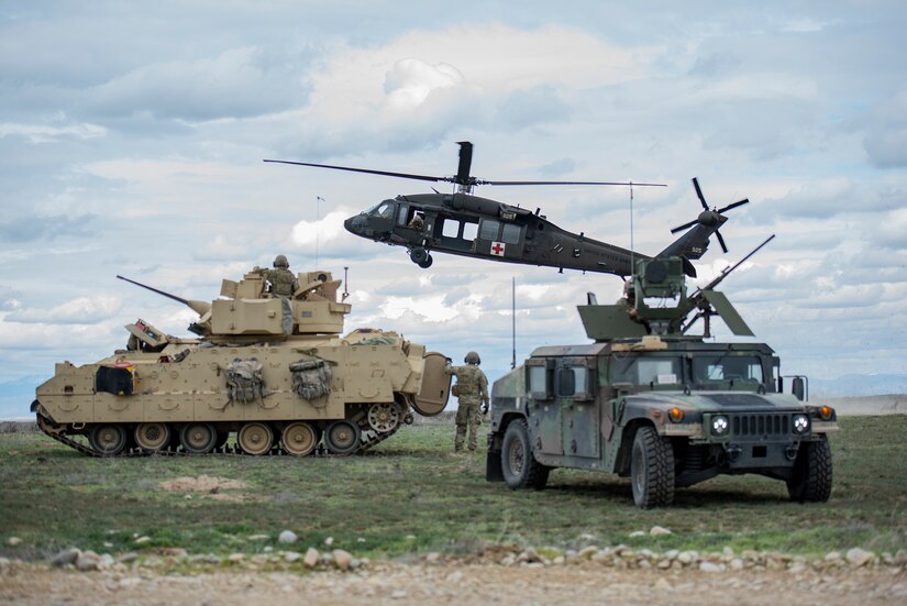 A soldier walks between a tank and a vehicle in a field as a helicopter hovers overhead.