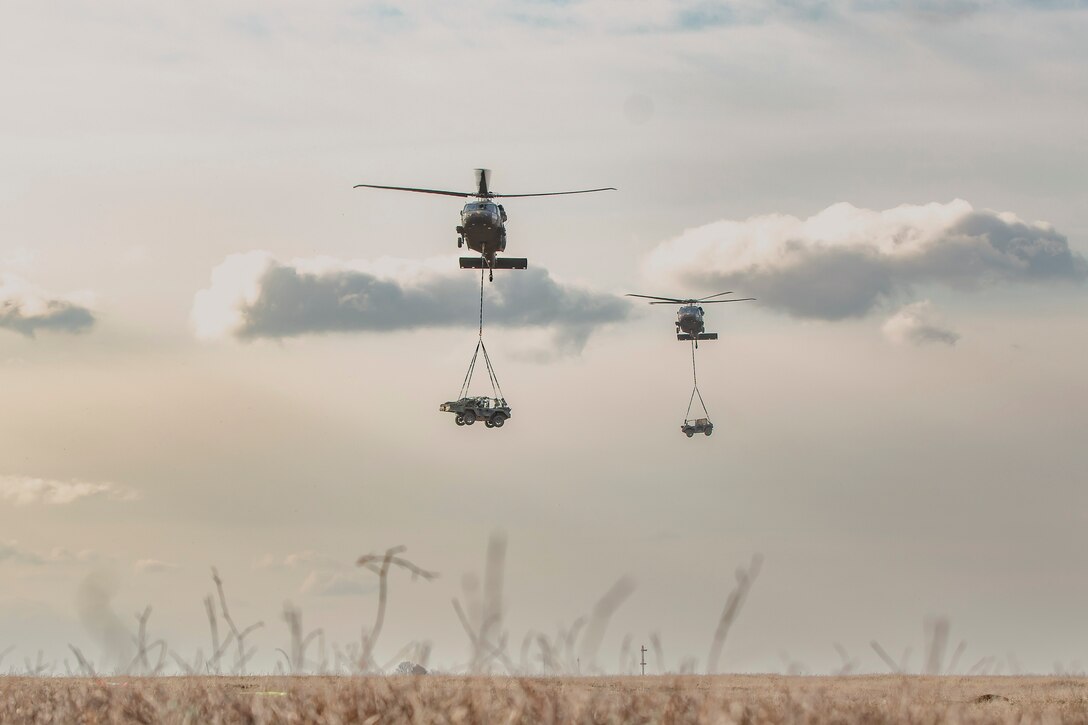 Two helicopters carrying vehicles fly next to each other over a field.