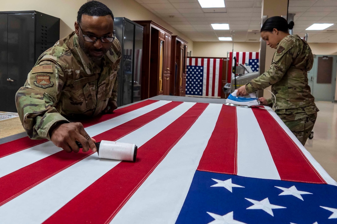 An airman uses a lint roller while another airman uses an iron on an American flag.