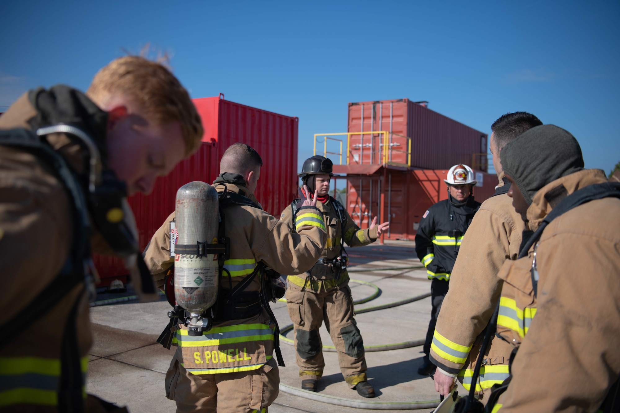 A group of firefighters speak in front of two red buildings.