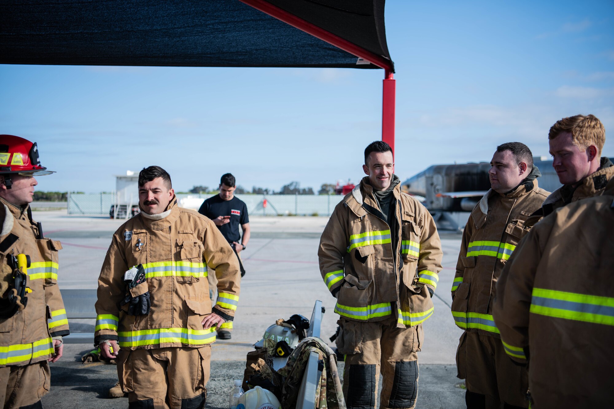 A group of firefighters in fire-protective gear stand under an awning.