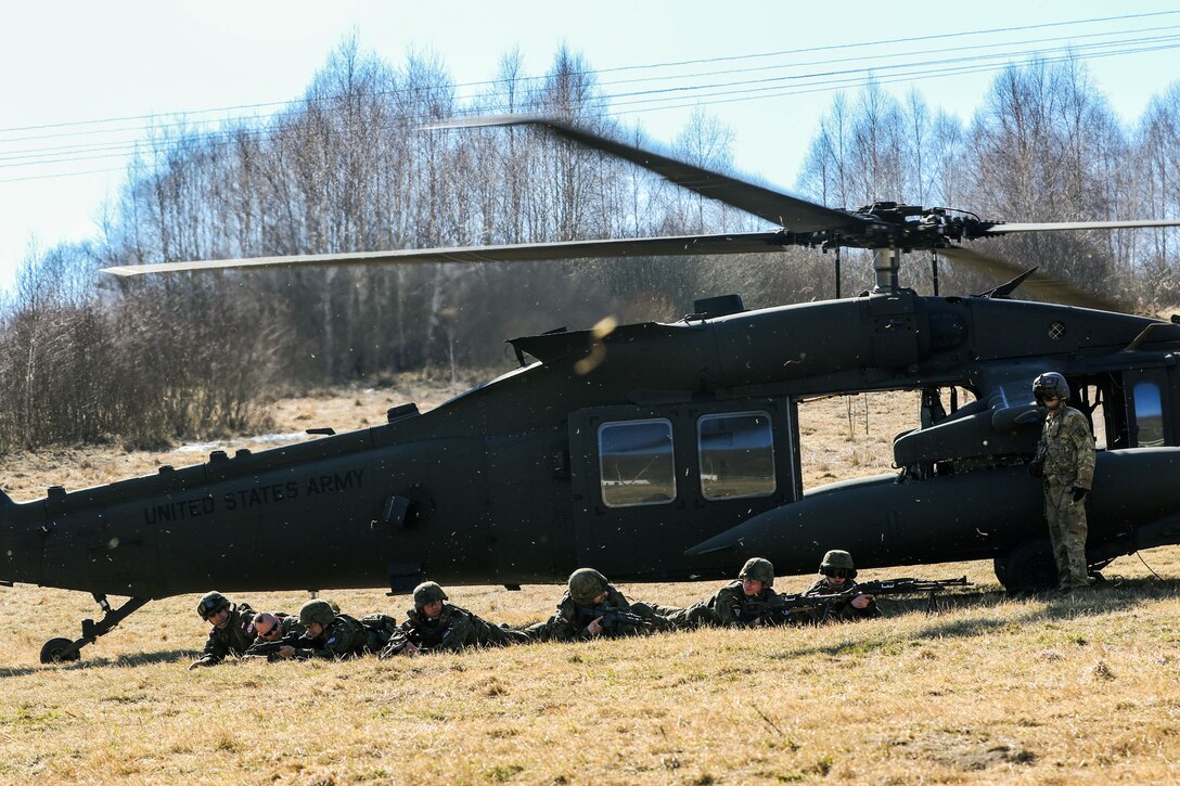 Polish soldiers lie prone in front of a helicopter parked in a field as U.S. soldier stands by.