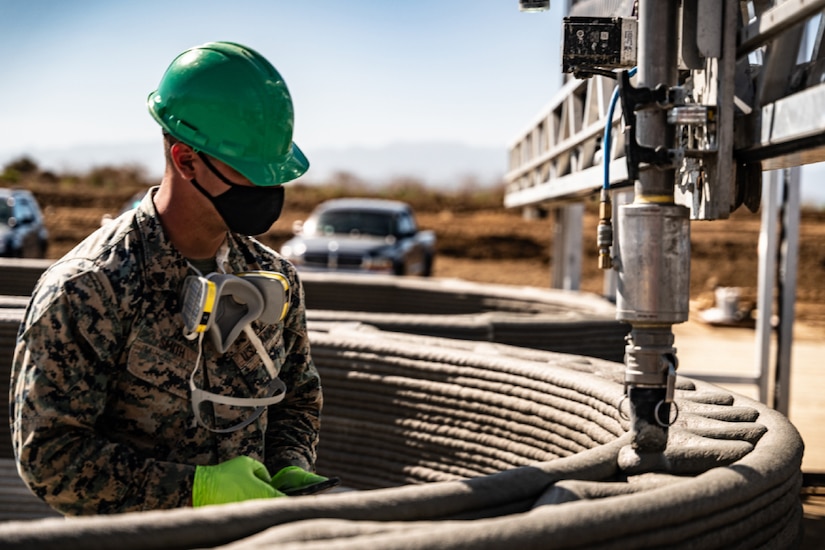 A uniformed Marine wearing construction gear monitors a concrete-laying machine.