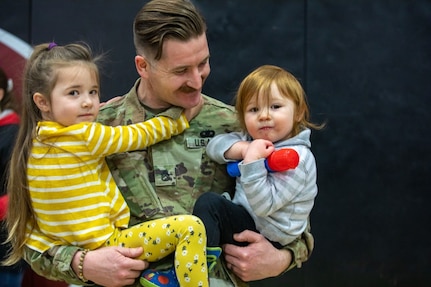 Staff Sgt. Anthony Streck, a supply sergeant with Headquarters and Headquarters Battery, 197th Field Artillery Brigade, holds two of his children, (from left to right) Vaira and Rowan, after a deployment ceremony April 2, 2022, at Concord High School.