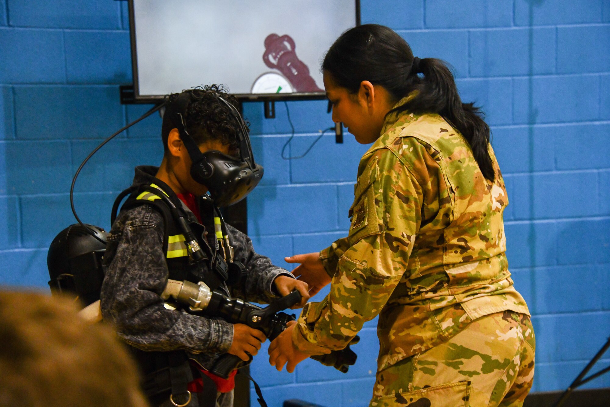 Airman 1st Class Gabriella Lopez, 97th Civil Engineer Squadron firefighter, helps an Altus Junior High School Student operate the FLAIM trainer at Altus High School, Altus, Oklahoma, March 30, 2022. The FLAIM Trainer simulates fires and other hazardous scenarios virtually to help firemen prepare for real-life events. (U.S. Air Force photo by Airman 1st Class Miyah Gray)