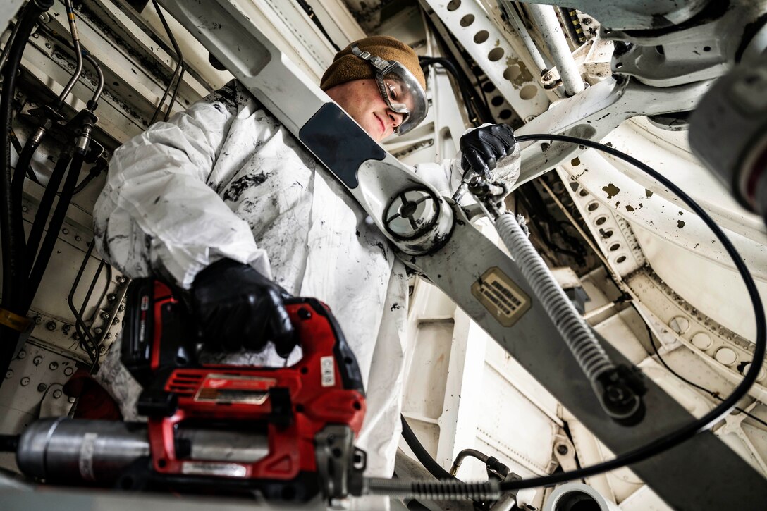 An airman wearing protective gear puts lubricant on an aircraft.